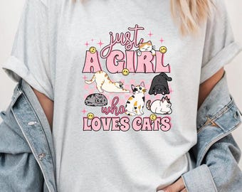 May include: Light gray t-shirt with the text "just a GIRL who LOVES CATS" in pink and black. The shirt features illustrations of several cats in various poses, along with smiley face graphics.