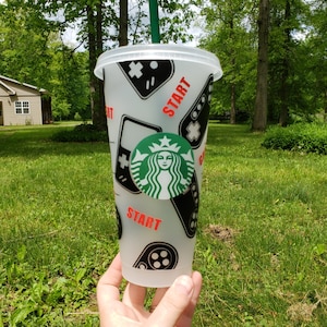 May include: A white reusable cup with a black and green design featuring a video game controller, the word "START" and the Starbucks logo.
