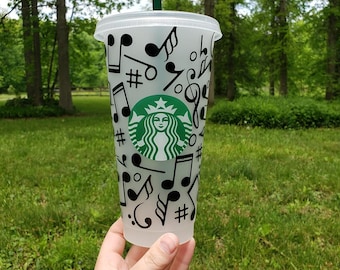 Peut inclure: Un gobelet réutilisable en plastique transparent avec un motif de notes de musique noires et blanches. Le gobelet porte le logo Starbucks sur le devant. Le gobelet est tenu par une main.