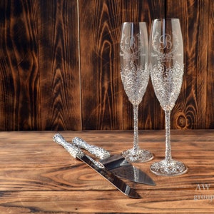 Op de afbeelding: Twee kristallen champagnefluiten met een bloemmotief en gepersonaliseerde initialen, en een zilveren taartmes en -serverset op een houten oppervlak.