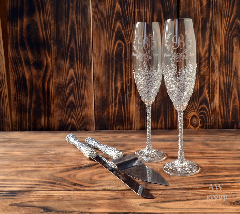 Pode incluir: Duas ta&ccedil;as de champanhe de cristal com um design floral e iniciais personalizadas, e um conjunto de faca e servidor de bolo de prata sobre uma superf&iacute;cie de madeira.