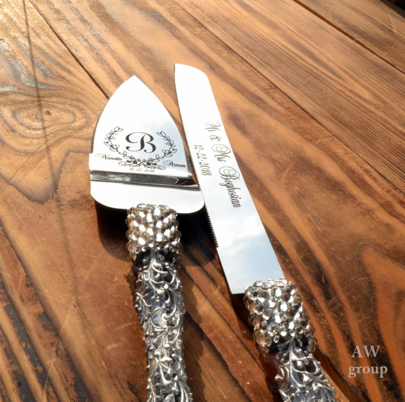 Pode incluir: Um conjunto de faca para cortar bolo de prata com uma pega decorativa e strass. A faca tem o texto "Mr. & Mrs. Beglarian 12.22.2018" gravado. O servidor tem o texto "Nanette & Armen 12.22.2018" e uma letra "B" em uma coroa gravada.
