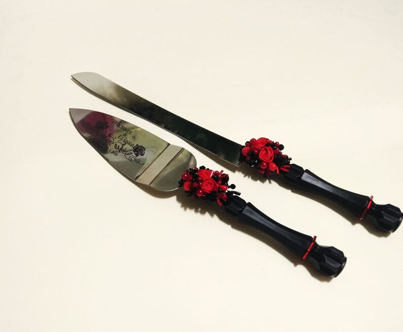 K&ouml;nnte beinhalten: Ein schwarzes und rotes Hochzeitstortenmesser-Set mit einem dekorativen Griff und dem eingravierten Text "Mr. & Mrs." auf der Klinge.