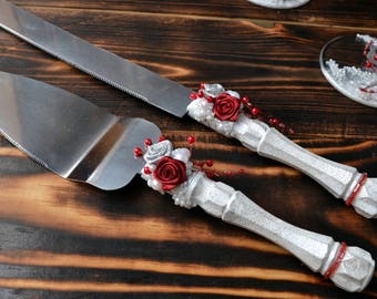 Könnte beinhalten: Ein Set aus zwei silbernen Kuchenschneidemessern mit weißen Griffen, die mit roten Rosen und perlmuttartigen Perlen verziert sind. Die Messer liegen auf einer Holzoberfläche.