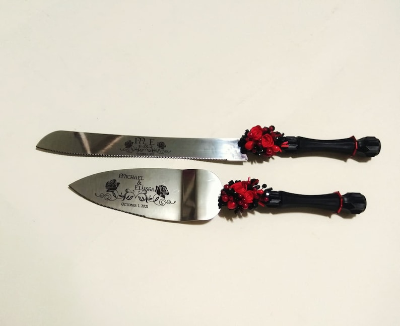 K&ouml;nnte beinhalten: Ein Set aus zwei Kuchenspachteln mit schwarzen Griffen und roten Rosenverzierungen. Das gr&ouml;&szlig;ere Messer ist mit "M.E." graviert und das kleinere Messer ist mit "Michael & Elyssa October 1, 2021" graviert.