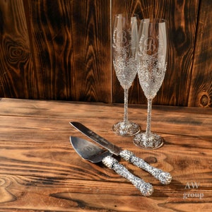 Pode incluir: Um conjunto de duas ta&ccedil;as de champanhe prateadas com um design decorativo e uma inicial gravada em cada copo. As ta&ccedil;as est&atilde;o sobre uma superf&iacute;cie de madeira ao lado de uma faca de bolo e um servidor prateados com um design decorativo.