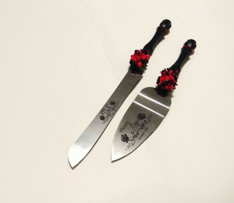 K&ouml;nnte beinhalten: Ein schwarzes und rotes Hochzeitstorten-Schneideset mit einer silbernen Klinge und einem schwarzen Griff, der mit roten Rosen verziert ist. Die Klinge ist mit dem Text "Mr. & Mrs. Michael & Eugenia October 1, 2016" graviert.