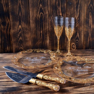 May include: Two clear glass champagne flutes with gold detailing and the text "Eric and Tina Anderson June 13, 2018" etched on the side. Two clear glass cake plates with gold detailing and the same text etched on the side. A gold cake knife with a silver blade.