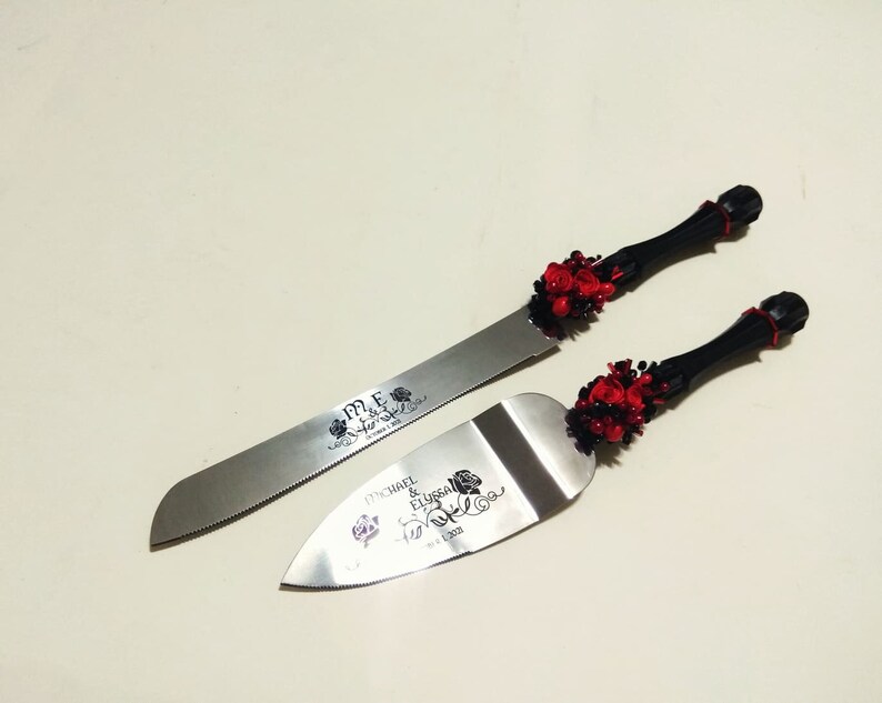 K&ouml;nnte beinhalten: Ein schwarzes und rotes Hochzeitstorten-Schneideset mit einer silbernen Klinge und einem schwarzen Griff, der mit roten Rosen verziert ist. Die Klinge ist mit "Mr. & Mrs." und "Michael & Elissa" sowie dem Datum "Oct. 2020" graviert.
