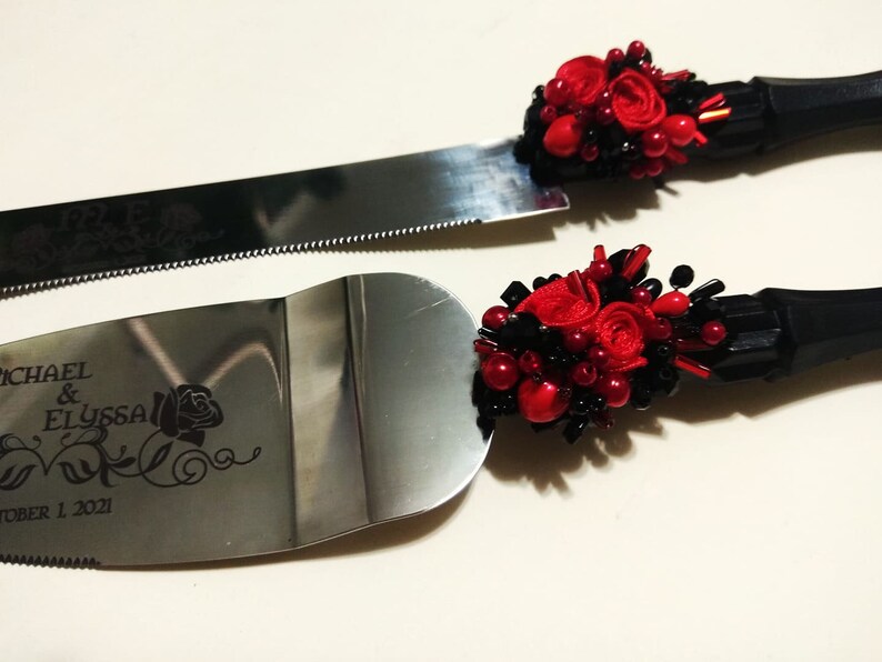 K&ouml;nnte beinhalten: Ein Set aus zwei silbernen Kuchenspachteln mit schwarzen Griffen und roten und schwarzen floralen Verzierungen. Die Spachteln sind mit dem Text "MICHAEL & ELYSSA OCTOBER 1, 2021" graviert.