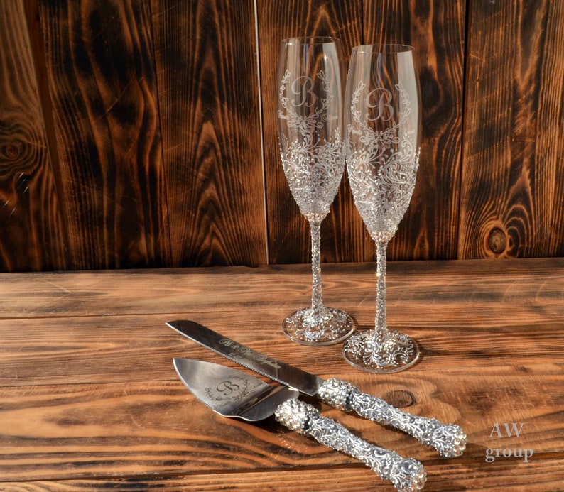 Pode incluir: Duas ta&ccedil;as de champanhe prateadas com um design floral e a letra "B" gravada na frente. Uma faca de bolo prateada com um design floral e uma al&ccedil;a de strass.