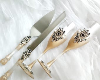 May include: A cake knife and server set alongside two champagne flutes. Each piece features a gold-toned ombre finish on the handles and stems, embellished with clusters of clear and black faceted crystals in an oval, eye-like arrangement.