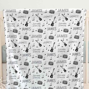 May include: A white minky fleece baby blanket with a black and white print of musical notes, guitars, and boomboxes. The blanket is personalized with the name "JAMES DAVIS" repeated throughout the design. The blanket measures 30 inches by 40 inches.