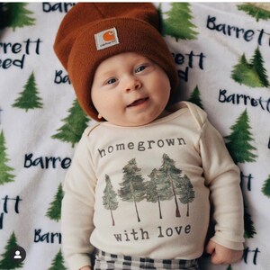 May include: A baby wearing a white long-sleeved onesie with the text "homegrown with love" and a picture of trees. The baby is also wearing a brown beanie and plaid pants.