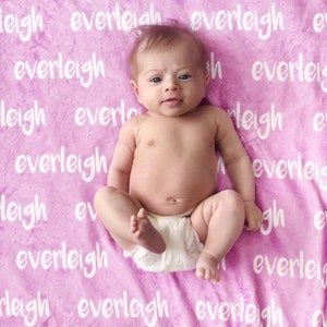 May include: A baby wearing a diaper is lying on a pink blanket. The blanket has the name "everleigh" printed repeatedly in white. The baby is looking at the camera.