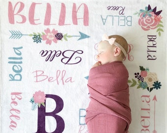 May include: A white baby blanket with a floral design and the name "Bella" repeated multiple times in different fonts and colors. The blanket also features a large purple "B" and a pink monogram. A baby is sleeping on the blanket.