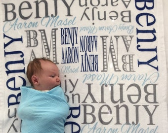 May include: A white baby blanket with the names Benjy, Aaron, and Masel printed in gray and blue lettering. A baby is swaddled in a light blue blanket and lying on the blanket.