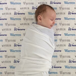 May include: A white swaddle blanket with a blue and gray pattern of the name "Warren" repeated across the fabric. A baby is swaddled in the blanket and is sleeping on the blanket.