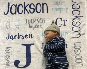 May include: A newborn baby wearing a navy blue and white striped onesie and a gray knotted hat, lying on a white blanket. The blanket is printed with the name "Jackson" in various fonts and colors.