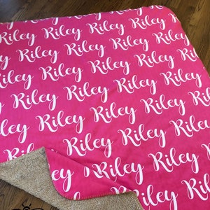 May include: A pink fleece blanket with the name "Riley" repeated in white cursive lettering. The blanket has a tan sherpa fleece backing. The text "Tan Sherpa Fleece" and "Stitched edge 50x60" and 60x80" is printed below the blanket.