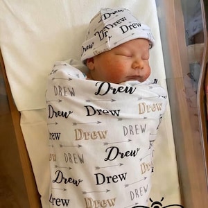 May include: A white swaddle blanket with the name "Drew" printed on it in black and gray. The blanket is wrapped around a baby wearing a white hat with the name "Drew" printed on it in black and gray. The baby is sleeping in a bassinet. The text "THE BEE BOUTIQUE NC, LLC" is printed on the blanket.