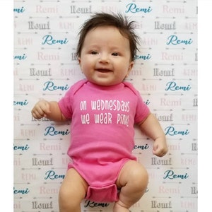 May include: A pink baby onesie with the text "on wednesdays we wear pink!" printed on the front. The onesie is worn by a baby lying on a white background with a repeating pattern of the name "Remi" in pink and blue.