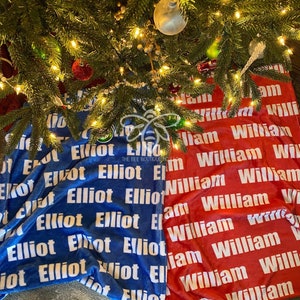 May include: A red and blue personalized Christmas tree skirt with the names "Elliot" and "William" repeated in white text.