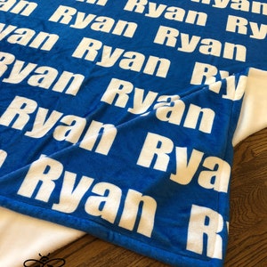 May include: A blue minky fleece blanket with the name "Ryan" printed in white all over the blanket. The blanket is folded in half and the text "Minky Fleece" is visible at the bottom of the image.