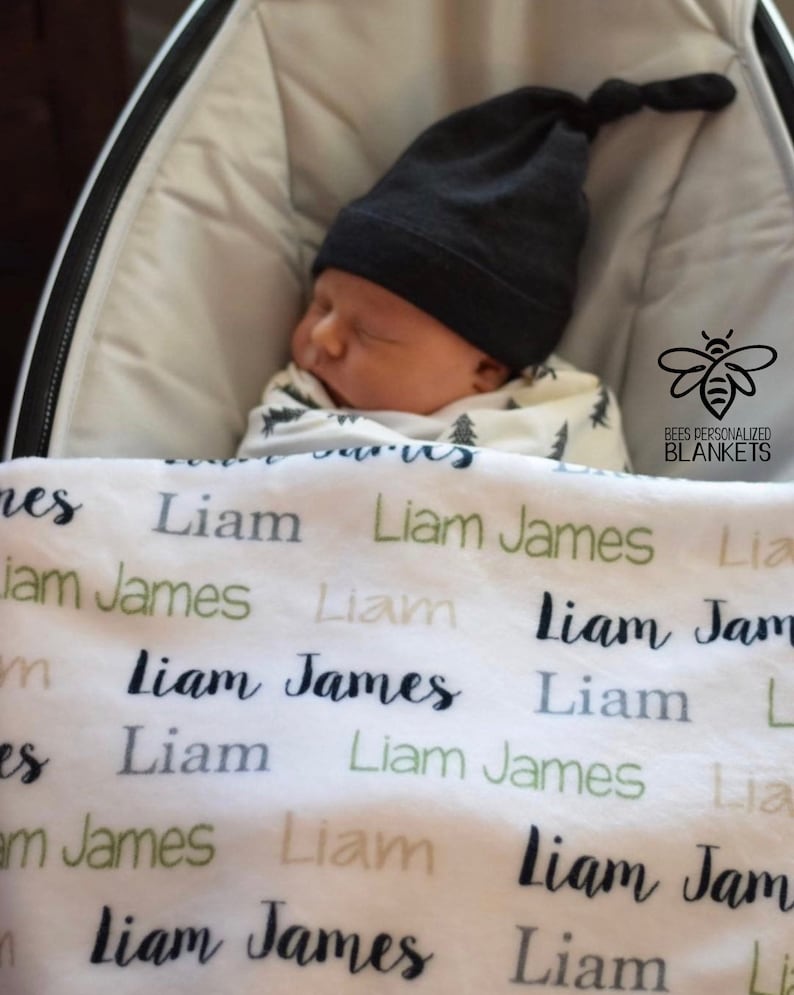 May include: A white baby blanket with the name "Liam James" repeated in black, green, and gray lettering. A baby is sleeping on the blanket wearing a black knit hat.