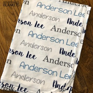 May include: White baby blanket with the name "Anderson Lee" repeated in blue and gray lettering. The blanket is made of thin jersey knit and has a folded edge.