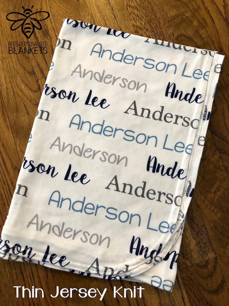 May include: White baby blanket with the name "Anderson Lee" repeated in blue and gray lettering. The blanket is made of thin jersey knit and has a folded edge.