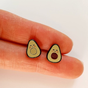 May include: A pair of avocado-shaped earrings with a cute cartoon face. The earrings are made of wood and have a brown seed in the center.