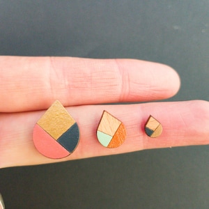 May include: Three wooden teardrop-shaped earrings in different sizes. The largest earring is painted pink, black, and gold. The medium earring is painted green, orange, and brown. The smallest earring is painted brown and gold.