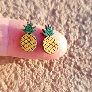 May include: Two wooden pineapple earrings. Each earring features a yellow pineapple body with a green top. The earrings are held between two fingers against a textured background.