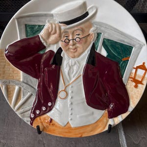 May include: A round ceramic decorative plate featuring a raised relief illustration of a person wearing a white top hat, a maroon jacket, a white vest, and round spectacles. The figure holds the brim of the hat while wearing a gold pocket watch chain.