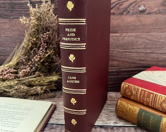 May include: A burgundy-coloured hardcover book titled "Pride and Prejudice" by Jane Austen. The spine features gold lettering and decorative accents. The book is standing upright, showcasing its elegant design.