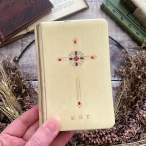 May include: A cream-colored rectangular book with a textured surface featuring a central cross design in red and black enamel accents. The cover includes the gold-stamped initials M. G. T. at the bottom and a vertical gold-toned spine edge.