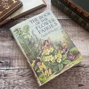 May include: A vintage copy of "The Book of the Flower Fairies" by Cicely M. Barker. The cover shows illustrations of fairies amongst flowers. The book is on a wooden surface with other antique books in the background.