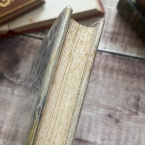 May include: Close-up of a vintage book with a worn, off-white cover and aged pages. The book's spine is visible, showing the title and author's name. The edges of the pages are yellowed, indicating age. The book is set against a wooden background.