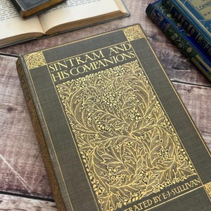 Pode incluir: Um close-up de um livro vintage intitulado "Sintram and His Companions" com letras douradas e um design floral decorativo na capa. O livro é ilustrado por E.J. Sullivan. Outros livros são visíveis no fundo.