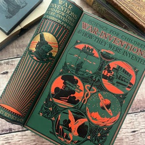 May include: An open vintage book titled "War Inventions and How They Were Invented" with a green cover and orange illustrations. The book's cover features a detailed illustration of a machine. The book is on a wooden surface.