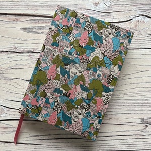 May include: A rectangular notebook with a fabric cover featuring a dense, repeating pattern of houses, trees, and foliage in shades of pink, blue, green, and black. A solid pink ribbon bookmark extends from the bottom edge of the notebook cover.