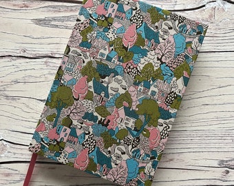 May include: A rectangular notebook with a fabric cover featuring a dense, repeating pattern of houses, trees, and foliage in shades of pink, blue, green, and black. A solid pink ribbon bookmark extends from the bottom edge of the notebook cover.