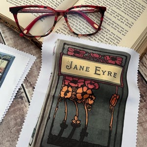 May include: A pair of red and black patterned eyeglasses rests on an open book. A scarf featuring the title "Jane Eyre" with floral illustrations is visible. The scarf has a gray background with red and orange flowers.