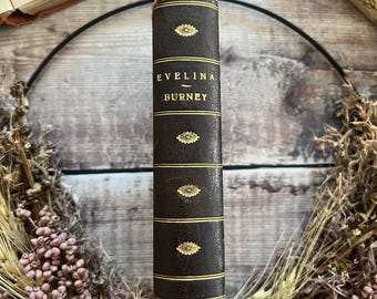 Puede incluir: Un lomo de libro encuadernado en cuero marrón oscuro con líneas horizontales y adornos florales dorados. El texto en el lomo dice Evelina de Burney en letras doradas. El libro está colocado verticalmente, mostrando su superficie texturizada.