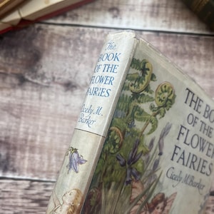 May include: A well-worn copy of "The Book of the Flower Fairies" by Cicely M. Barker. The cover is adorned with floral and fairy illustrations. The spine shows the title in blue lettering.