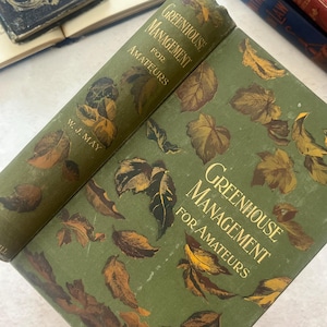 以下が含まれることがあります： W. J. May著のハードカバー本『Greenhouse Management for Amateurs』。表紙はオリーブグリーンで、茶色と金色の秋の葉の模様が繰り返されています。タイトルは表紙と背表紙の両方に金色の文字で印刷されています。