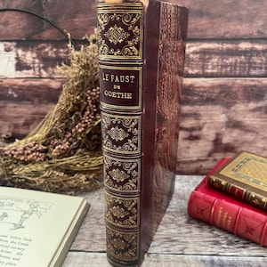 May include: A vintage book titled "LE FAUST DE GOETHE" with a burgundy leather cover and gold embossed detailing. The spine features decorative patterns and the title in gold lettering. Other books are visible in the background.