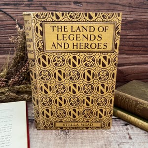 以下が含まれることがあります： ステラ・ミード著「The Land of Legends and Heroes」というタイトルのヴィンテージ本。茶色の「N」のデザインが繰り返されるパターンが特徴のタン色の表紙。タイトルは茶色の装飾的なフォントです。本の背表紙が見え、背景には他の本があります。