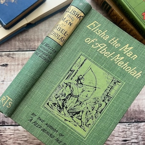 Puede incluir: Un libro antiguo titulado "Elisha the Man of Abel-Meholah" con una cubierta verde y letras doradas. La portada presenta una ilustración de un hombre con arco y flecha. El lomo también es verde con una etiqueta de título.